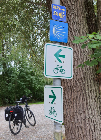 im Vordergrund Richtungspfeile für Radweg, darüber das Muschelzeichen für den Jakobsweg, im Hintergrund Susas Fahrrad mit 2 Packtaschen