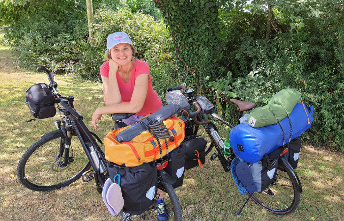 Susa Hofmeister mit Käppi und pinkem T-Shirt neben ihrem Fahrrad, das mit mehreren Taschen bepackt ist, daneben das ebenfalls schwer bepackte Fahrrad ihres Mannes