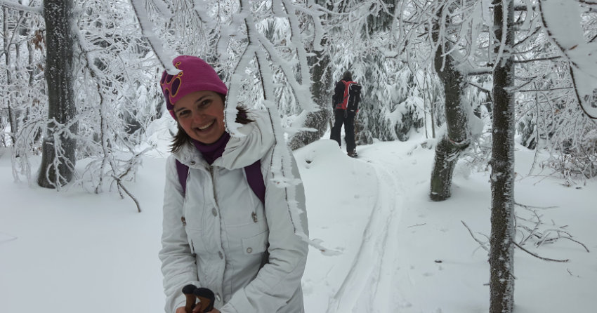 Susa Hofmeister lachend mit weißer Winterjacke, pinker Mütze und Rucksack in einem stark verschneiten Waldstück mit Schneeschuhen und Stöcken