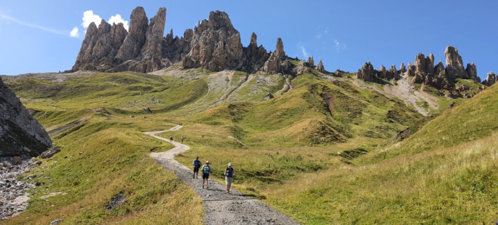 Im Vordergrund grüne Hügel und dazwischen ein sich windender Kiesweg, auf dem mehrere Wanderer unterwegs sind. Im Hintergrund eine Felsformation aus hohen, spitzen bräunlichen Felsen.